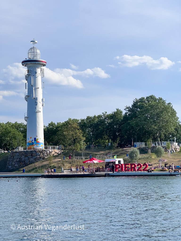 Ein weißer Aussichtsturm mit Blick auf die Donau, an dem das große rote „PIER22“-Schild prangt, und darunter ein belebter Treffpunkt am Ufer. Die Leute entspannen sich am Flussufer, während Bäume und der blaue Himmel den modernen Erholungsbereich einrahmen.