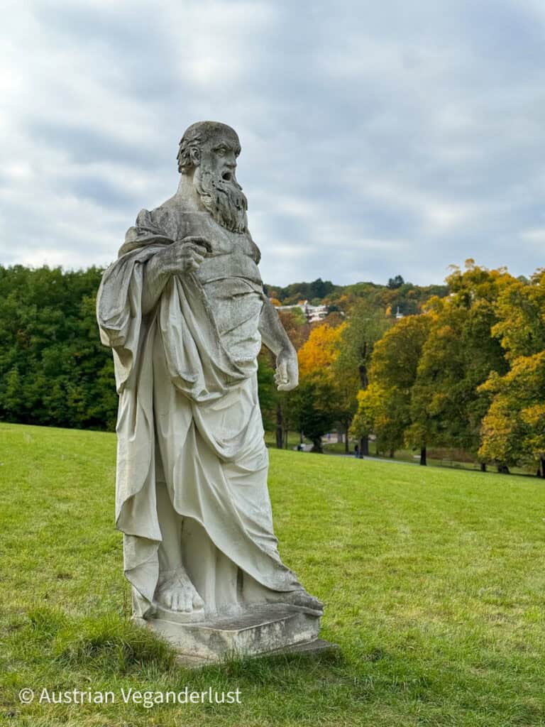 Eine verwitterte Steinstatue einer bärtigen, in eine Robe gekleideten Gestalt steht auf einem grasbewachsenen Hügel und blickt auf einen Park voller herbstlich gefärbter Bäume. Sanfte graue Wolken ziehen über den Himmel und bilden eine dramatische Kulisse für die historische Skulptur.