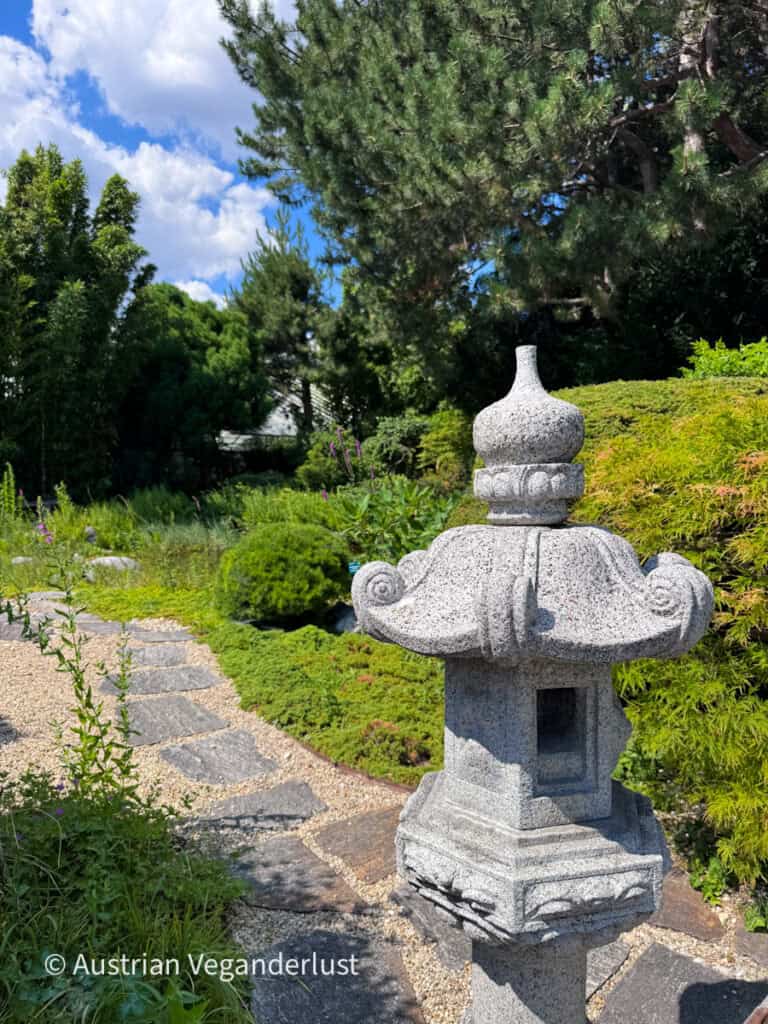 Eine traditionelle Steinlaterne steht neben einem gewundenen Steinweg in einem japanischen Garten, umgeben von gepflegten Sträuchern, immergrünen Bäumen und üppigem Grün. Ein strahlend blauer Himmel und sanfte weiße Wolken rahmen die friedliche Gartenlandschaft ein.