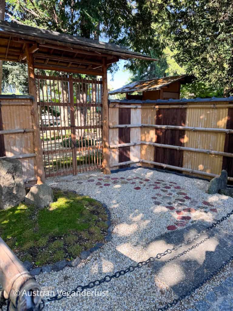Ein traditionelles Holztor und ein Bambuszaun markieren den Eingang zu einem japanischen Garten, der mit weißem Kies, roten Trittsteinen und großen Felsen in einem minimalistischen Design gestaltet ist. Sonnenlicht fällt durch die umliegenden Bäume und wirft sanfte Schatten über den Eingang.