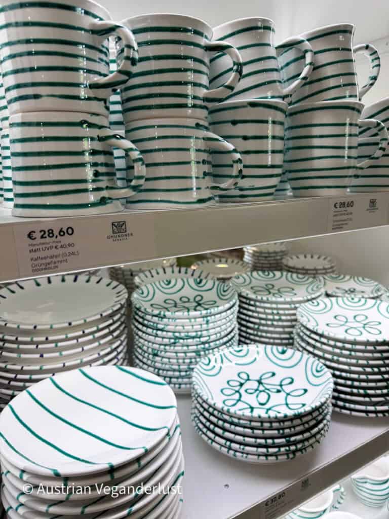 Shelves of Gmundner Keramik pottery with white ceramic mugs and plates decorated in hand painted green stripes and looping patterns. Price labels show mugs at "€ 28.60" and plates at "€ 32.80."