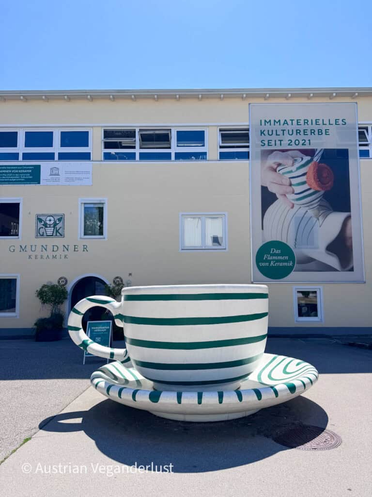 Giant white and green striped ceramic cup and saucer outside the Gmundner Keramik building. A large sign on the building reads "IMMATERIELLES KULTURERBE SEIT 2021" and "Das Flammen von Keramik."