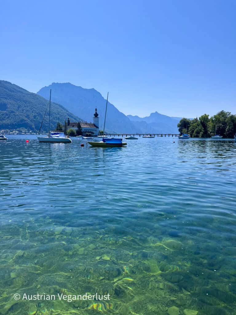 Sailboats floating on the clear blue water of Lake Traunsee with Schloss Ort and its long wooden bridge in the distance. Steep green mountains rise behind the castle under a cloudless blue sky.