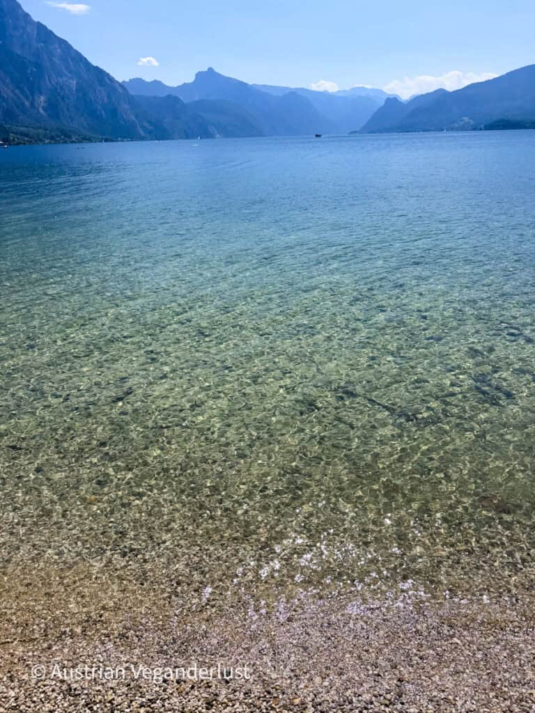 Exceptionally clear shallow water along the shore of Lake Traunsee with the pebbled lakebed visible beneath the surface. The blue lake stretches toward dramatic mountain ridges in the distance.