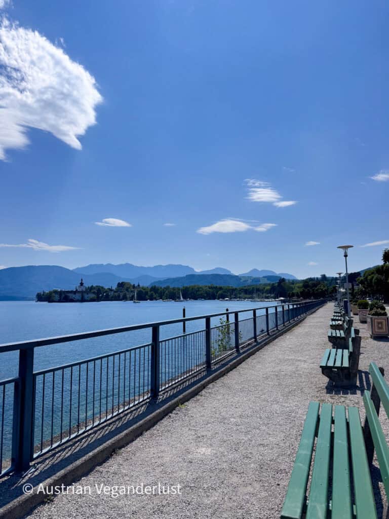 Wide lakeside promenade in Gmunden with green benches overlooking Lake Traunsee. Schloss Ort appears across the blue water with layers of mountains beyond it.