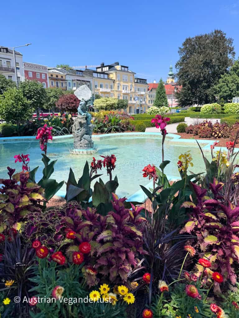 Colorful flower beds surrounding a pale blue fountain in a landscaped garden in Gmunden. Historic pastel buildings and a church tower rise behind the garden under a clear blue sky.