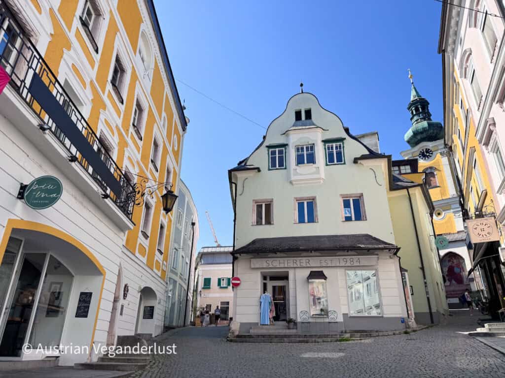 Narrow cobblestone street in Gmunden lined with pastel historic buildings and small shops. A yellow church tower with a green onion shaped dome rises between the buildings beneath a bright blue sky.