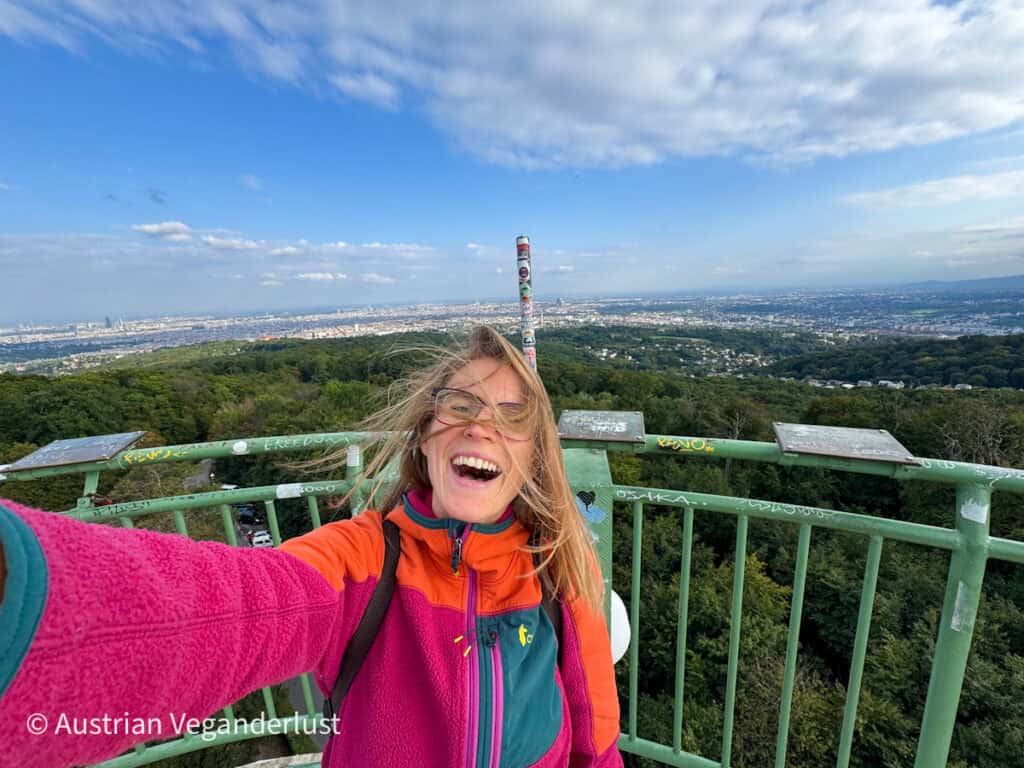 Eine lächelnde Frau macht ein Selfie im Wind auf der Aussichtsplattform der Jubiläumswarte, während weit unter ihr die Wälder und die weitläufige Stadt Wien zu sehen sind. Ihr Haar weht ihr ins Gesicht, während sie neben dem grünen Metallgeländer steht.
