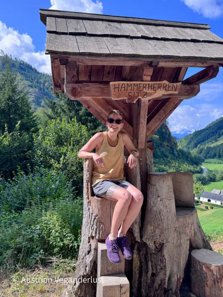 Tina in hiking clothes and sunglasses sitting on a rustic wooden seat overlooking a green mountain valley. A carved sign above her reads "HAMMERHERREN SITZ."