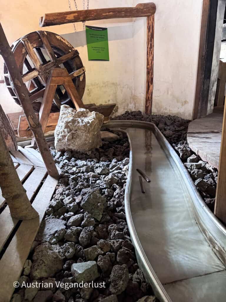 Historic water powered mechanism with a large wooden wheel beside a narrow water channel inside a rustic mill building. A sign reads "Das Wasserrad treibt den Hammer der Schmiede." and "The waterwheel boosts the hammer of the smith."