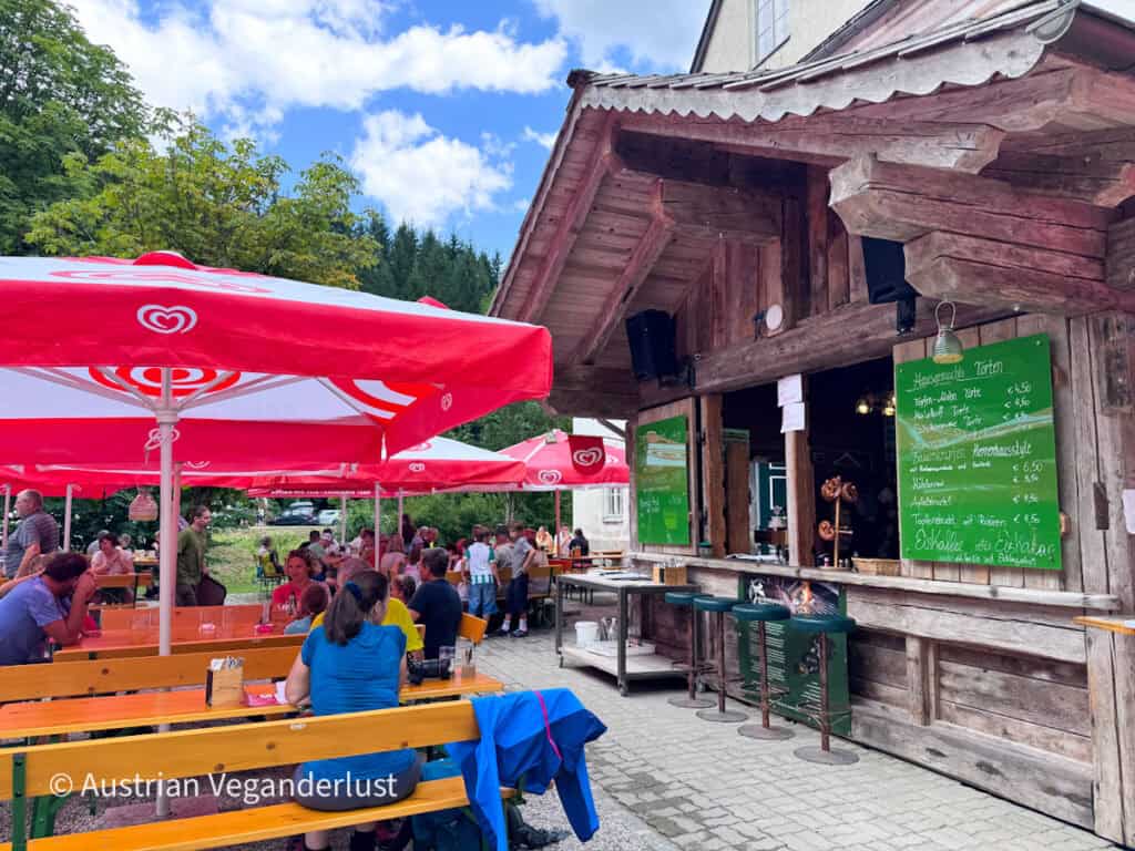 Busy outdoor restaurant with wooden picnic tables beneath red umbrellas beside a rustic timber building. Hikers and visitors sit outside for food and drinks surrounded by forest.