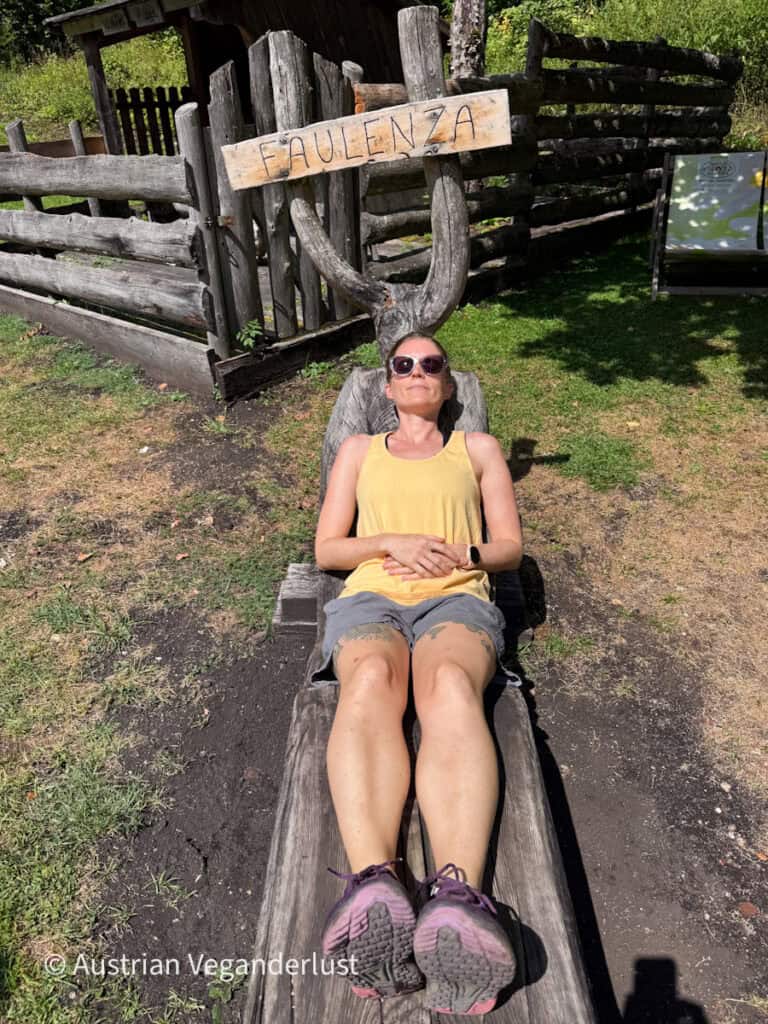 Tina in hiking clothes and sunglasses relaxes on a rustic wooden lounger in the sunshine. A handmade wooden sign above her reads "FAULENZA."