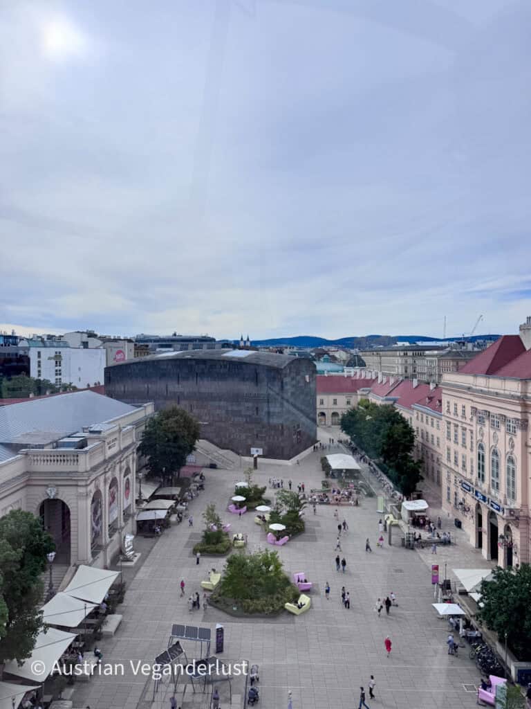 Blick über das MuseumsQuartier in Wien, wo Menschen im großen, fußgängerfreundlichen Innenhof zwischen historischen Gebäuden spazieren gehen und sich entspannen. Die dunkle Steinfassade des mumok ragt am anderen Ende des Platzes deutlich hervor.