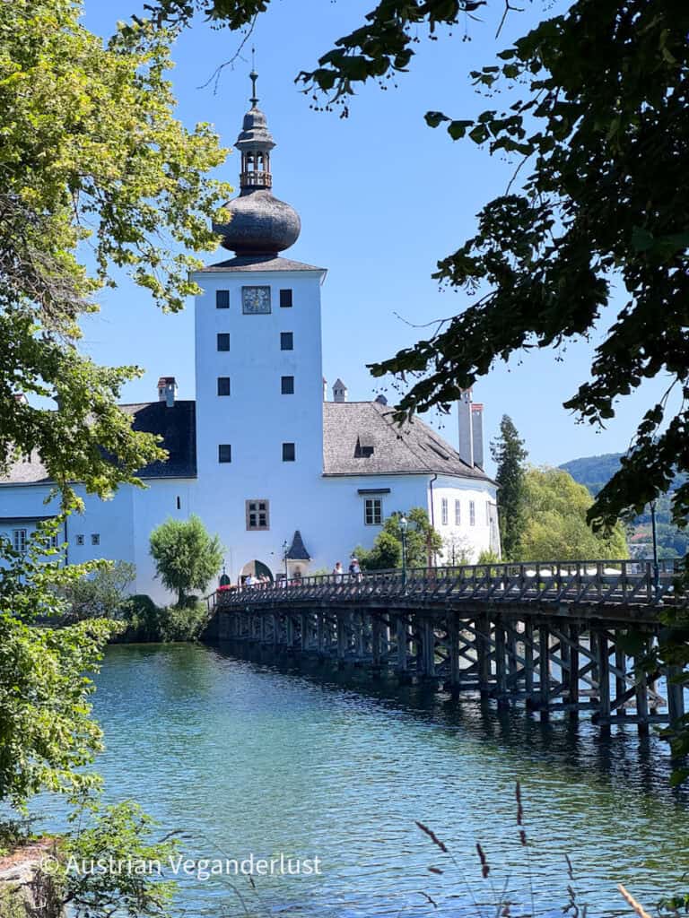 Schloss Ort rising from the blue water of Lake Traunsee with its white clock tower and dark onion shaped dome framed by leafy trees. A long wooden bridge carries visitors across the lake to the castle entrance.