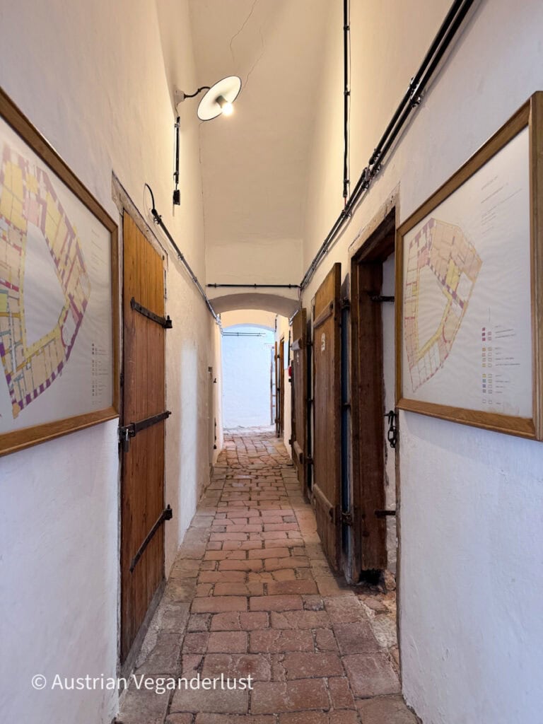 Narrow historic corridor with white plaster walls. Wooden doors line the brick floor walkway with framed maps hanging on both sides.