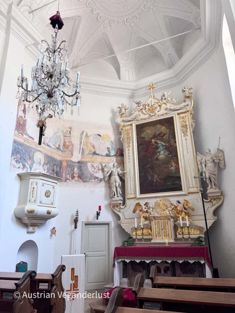 Ornate church interior with a crystal chandelier hanging beneath a decorative white vaulted ceiling. A gilded altar with angels and a large religious painting stands beside faded wall frescoes and a white pulpit.