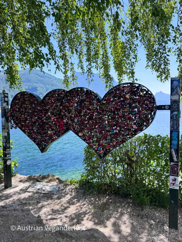Two large metal hearts densely covered in colorful love locks beside a lake. Leafy branches frame the hearts with blue water and forested mountains visible behind them.