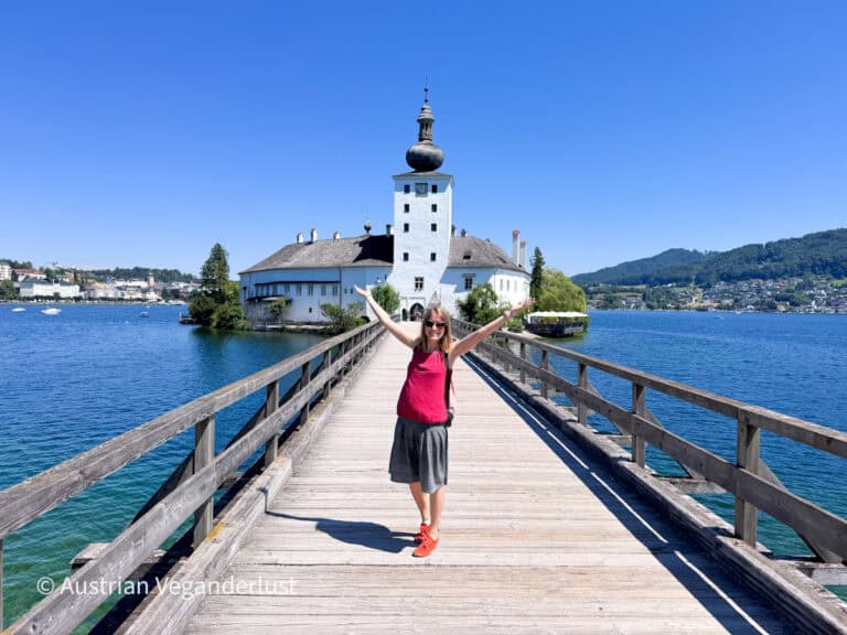 Tina in sunglasses standing on a long wooden bridge over a bright blue lake with both arms raised. The bridge leads directly to a white lakeside castle with a tall onion domed tower and mountains beyond.