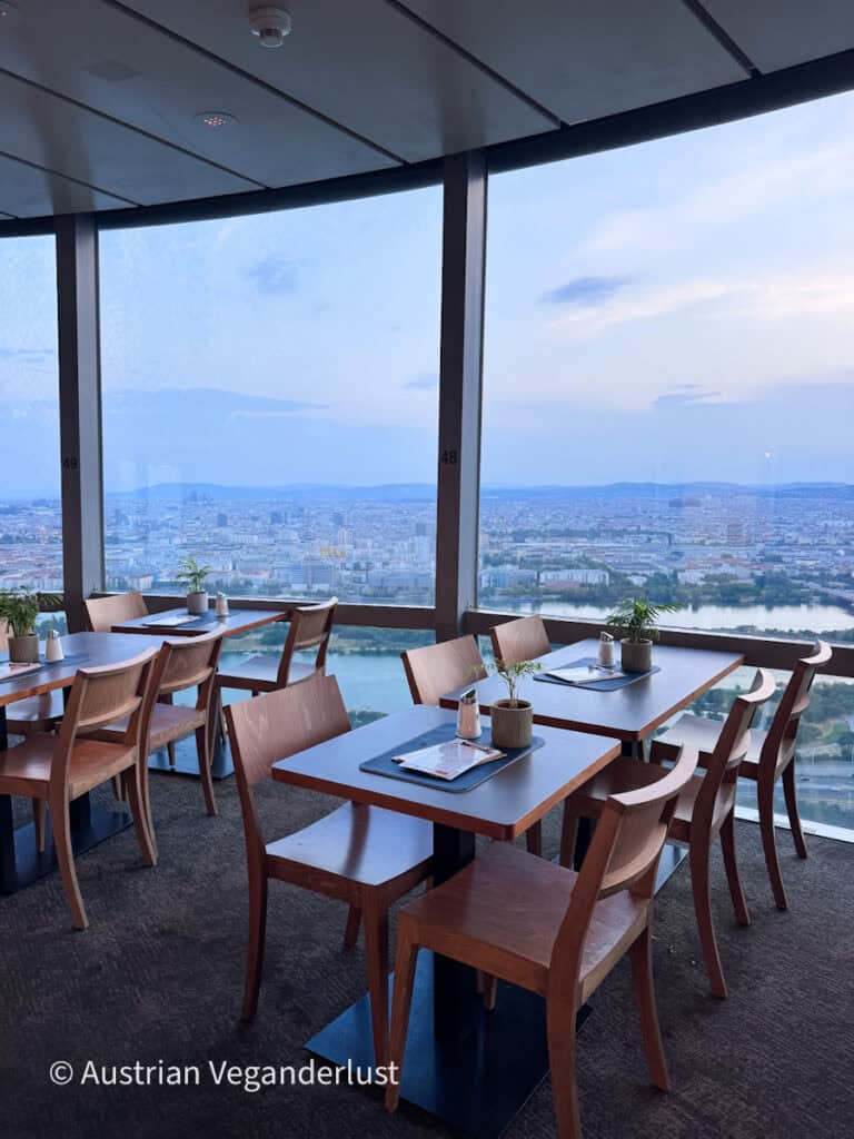 Restaurantbereich hoch über Wien mit Holztischen und -stühlen neben raumhohen Fenstern. Durch die Fenster hat man einen weiten Blick über die Stadt und die Donau.