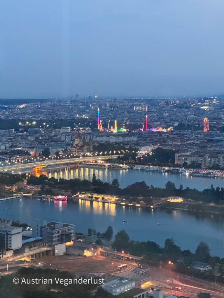 Wien bei Einbruch der Dämmerung, von hoch über der Stadt gesehen. Die Donau spiegelt die Lichter der Brücken und Gebäude wider, während in der Ferne die bunten Fahrgeschäfte des Vergnügungsparks leuchten.