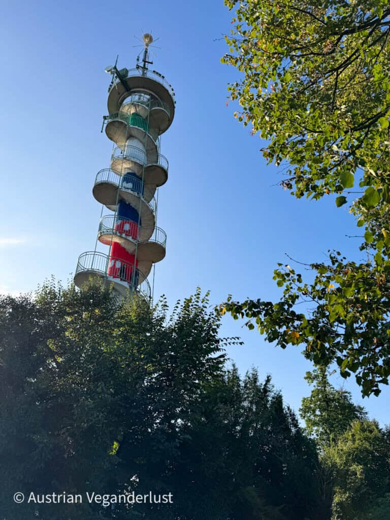 Der Aussichtsturm „Jubiläumswarte“ ragt vor dem Hintergrund klarer blauer Himmel über die grünen Bäume empor. Der Betonturm verfügt über eine offene Wendeltreppe, die sich um seine Außenseite windet.