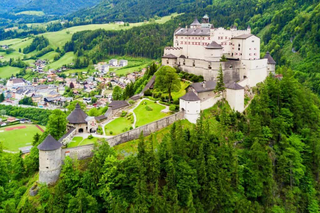 Die Burg Hohenwerfen thront hoch oben auf einem bewaldeten Hügel über einem kleinen österreichischen Ort. Die weitläufige weiße Festung hat Steinmauern. Runde Wehrtürme. Und dahinter erheben sich die Berghänge.