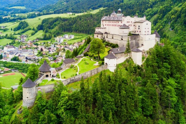 Die Burg Hohenwerfen thront hoch oben auf einem bewaldeten Hügel über einem kleinen österreichischen Ort. Die weitläufige weiße Festung hat Steinmauern. Runde Wehrtürme. Und dahinter erheben sich die Berghänge.