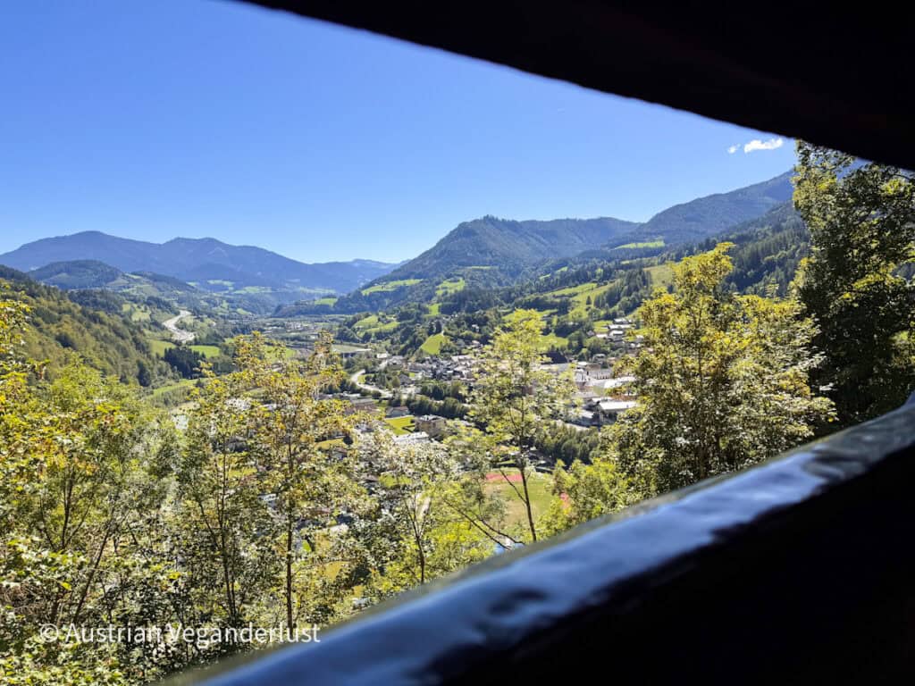 Blick durch eine dunkle Burgöffnung auf ein grünes Tal voller Häuser. Straßen. Wälder. Und sanfte Berge unter einem strahlend blauen Himmel.