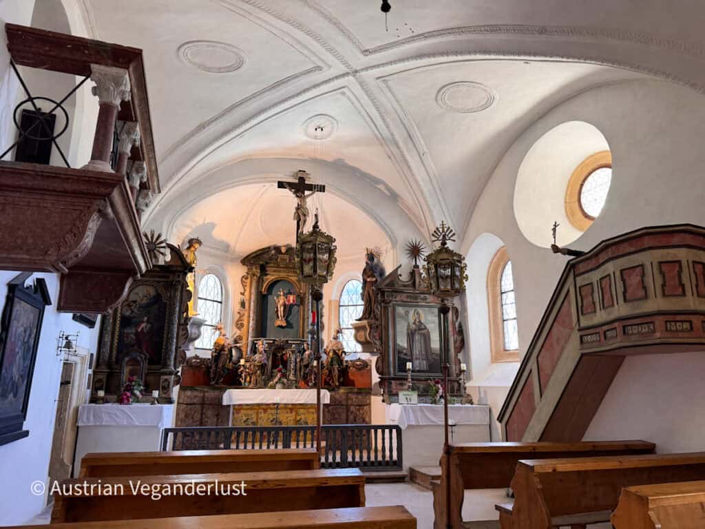 Das Innere einer kleinen Schlosskapelle mit hölzernen Kirchenbänken, die auf einen reich verzierten Altar ausgerichtet sind, der mit religiösen Gemälden und Statuen geschmückt ist. Weiße Gewölbedecken. Bogenfenster. Und eine verzierte Kanzel umgibt den Altarraum.