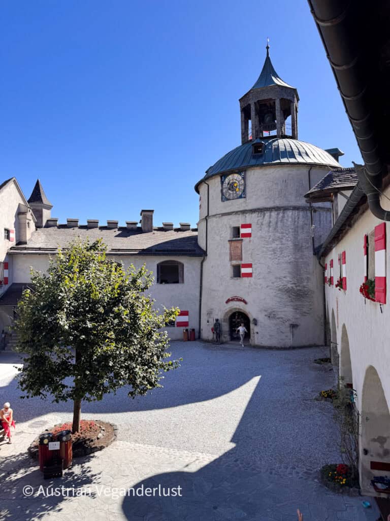 Ein sonniger Schlosshof, umgeben von weißen Gebäuden mit rot-weißen Fensterläden. Über dem Hof ragt ein runder Turm mit einer Uhr und einem offenen Glockenturm empor.