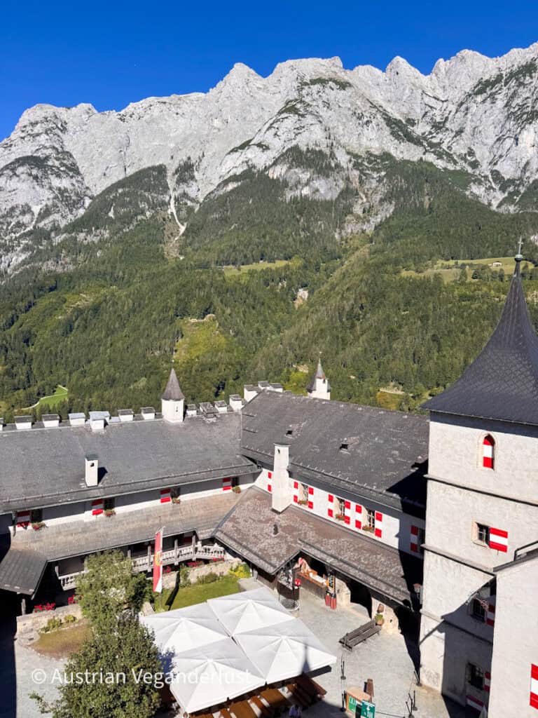 Blick über den Schlosshof hin zu den beeindruckenden hellgrauen Berggipfeln unter einem tiefblauen Himmel. Weiße Schlossgebäude mit rot-weißen Fensterläden umgeben unten Tische und Sonnenschirme.