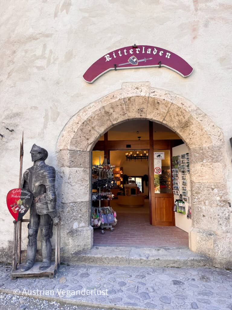 Ein gewölbter Steineingang zu einem Ritterladen, neben dem eine komplette mittelalterliche Rüstung steht. Auf einem burgunderroten Schild über dem Eingang steht „Ritterladen“.