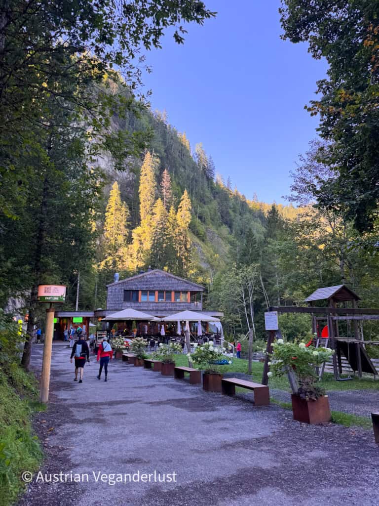 Wooden lodge and visitor area at the entrance to a forested gorge. People walk toward the building while steep cliffs and sunlit trees rise behind it.