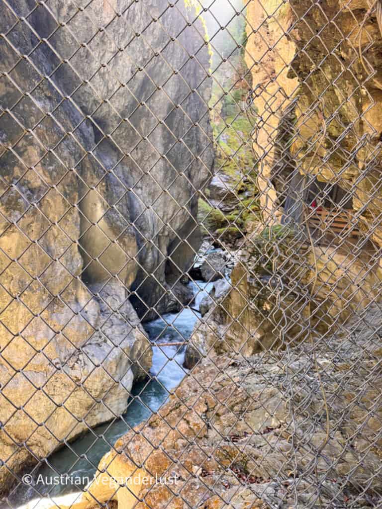 Fast flowing turquoise water winds through a deep rocky channel seen through protective wire mesh. Steep stone walls rise closely on both sides of the gorge.