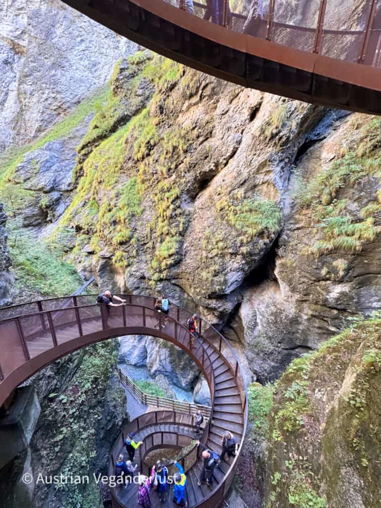 Visitors climb a winding metal staircase suspended between steep moss covered rock walls. Multiple levels of the walkway curve through the narrow gorge above the water.
