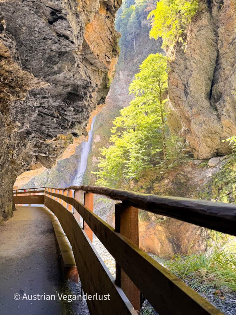 Ein Holzsteg führt entlang der Felswand durch die Schlucht, hinter dem Geländer ist ein hoher Wasserfall zu sehen. Zwischen den hoch aufragenden Felsen wachsen sonnenbeschienene grüne Bäume.