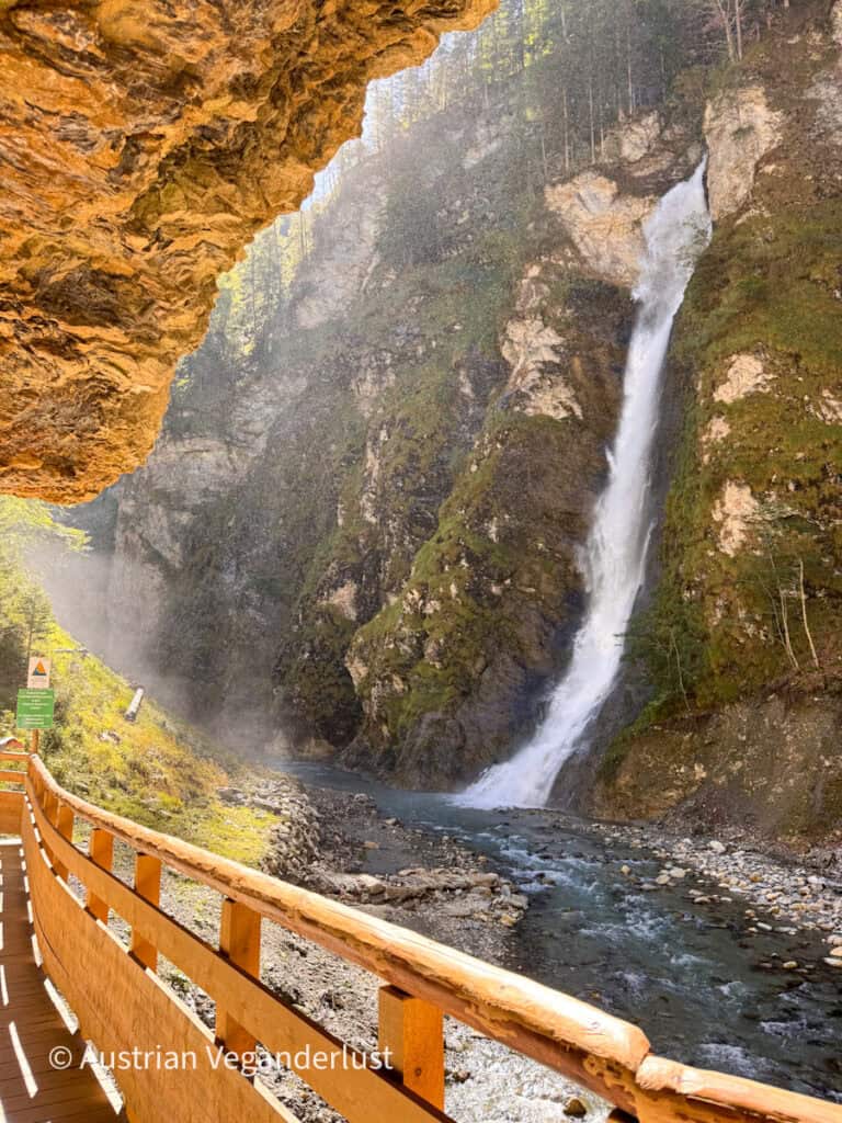 Tall waterfall plunges down a steep mossy cliff into a rocky stream beside a wooden gorge walkway. Mist rises around the falls in the sunlight.