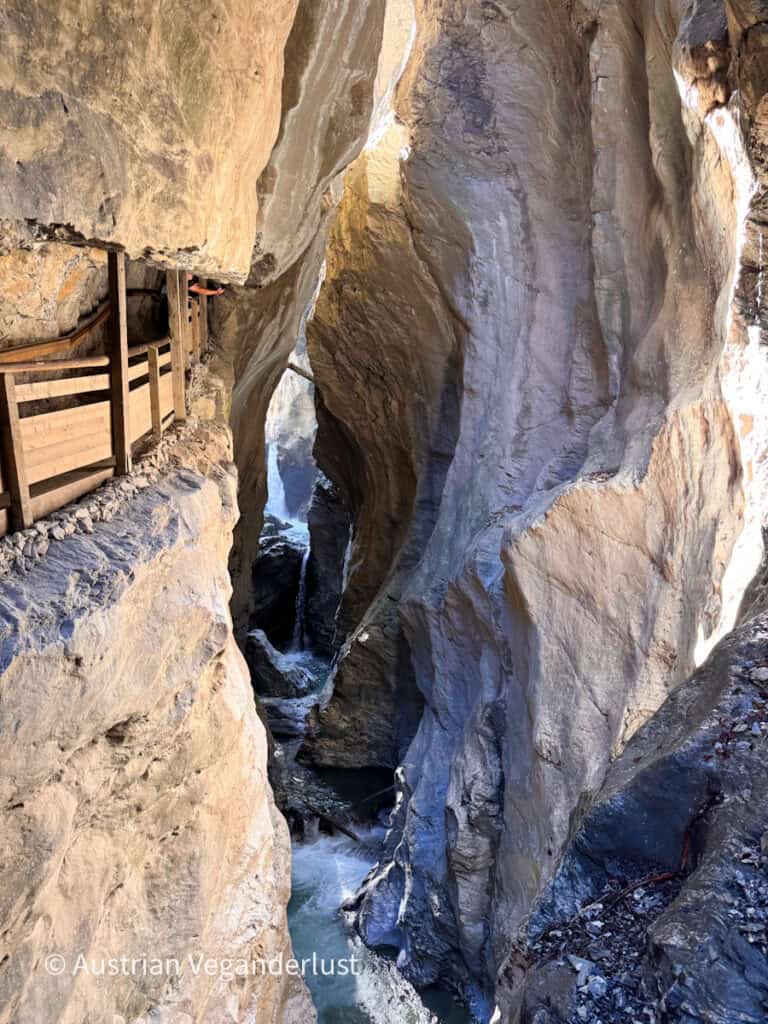 Narrow wooden walkway clings to a ledge high inside the gorge while water cascades through the deep rock channel below. Massive smooth stone walls surround the path and emphasize the depth and narrowness of the gorge.