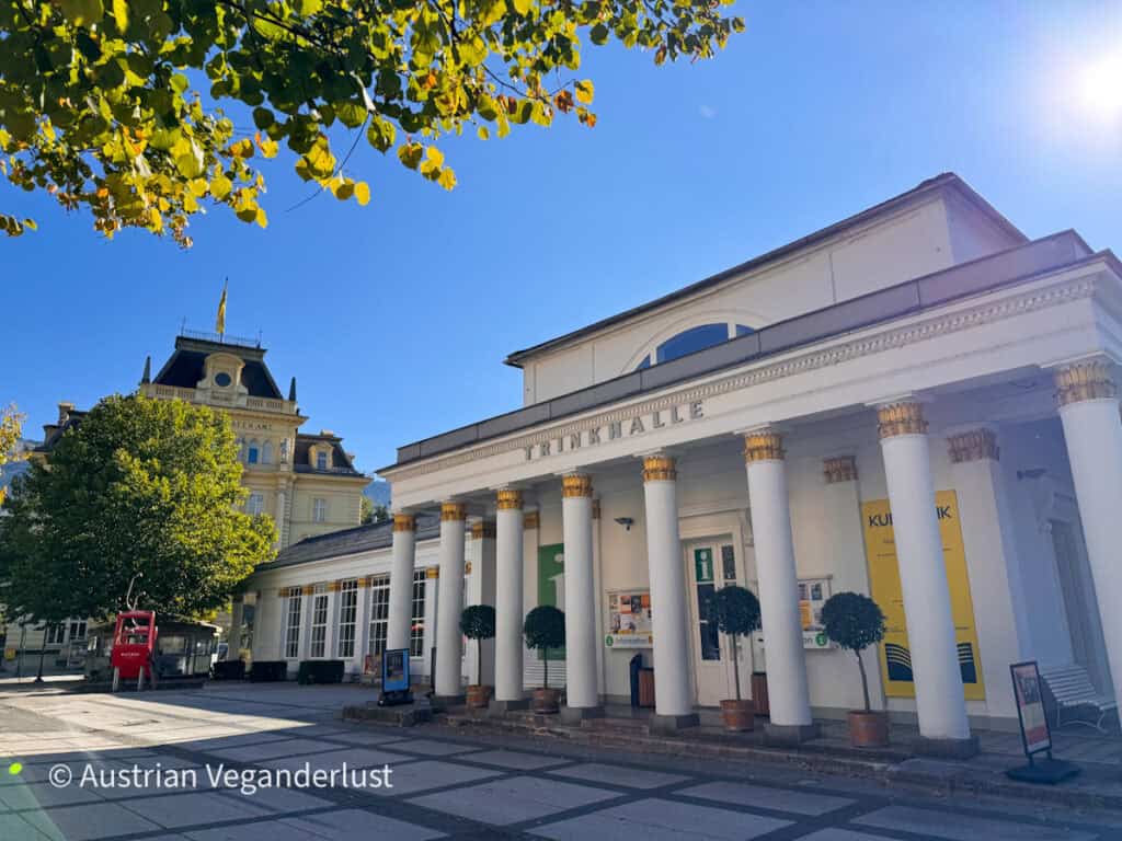 White neoclassical pavilion with gold topped columns and the word “TRINKHALLE” above the entrance. Small potted trees stand between the columns. A grand historic building with a flag on its roof rises behind it under a blue sky.