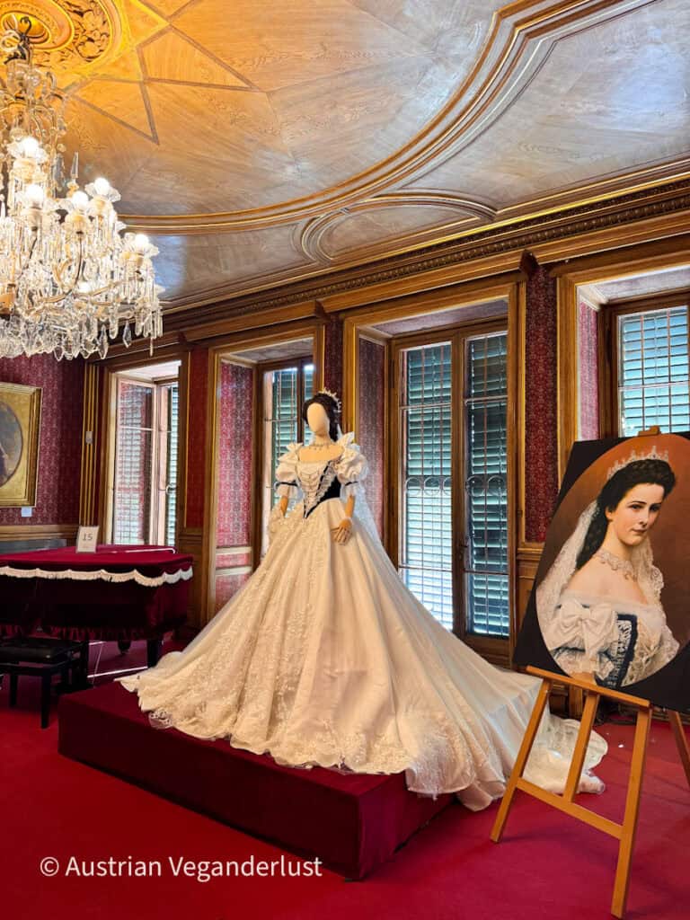 Replica of a white ball gown with puffed sleeves and a long train displayed on a red platform. A painted portrait of Empress Sisi with a tiara stands on an easel beside it. The room has a carved wooden ceiling and a crystal chandelier and red patterned walls.
