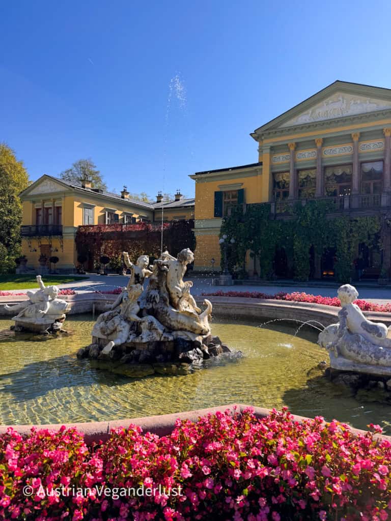 Marble fountain with cherub sculptures and a tall water jet in front of the yellow Kaiservilla in Bad Ischl. Bright pink flowers border the fountain basin. Ivy covers part of the villa under a clear blue sky.