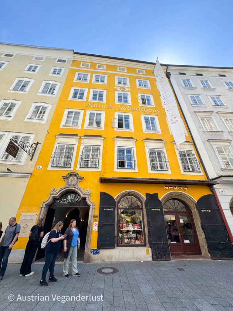 The bright yellow facade of "Mozarts Geburtshaus" in Salzburg, Austria, with ornate Baroque entrance and a SPAR grocery store on the ground floor. Several visitors stand on the cobblestone street in front of the historic building.