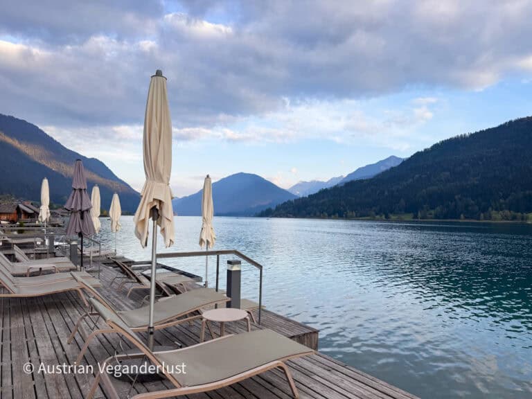 Wooden sun deck of Strandhotel Weissensee with loungers and closed beige umbrellas at the water’s edge. Calm turquoise lake water stretches between forested mountains under a soft evening sky.