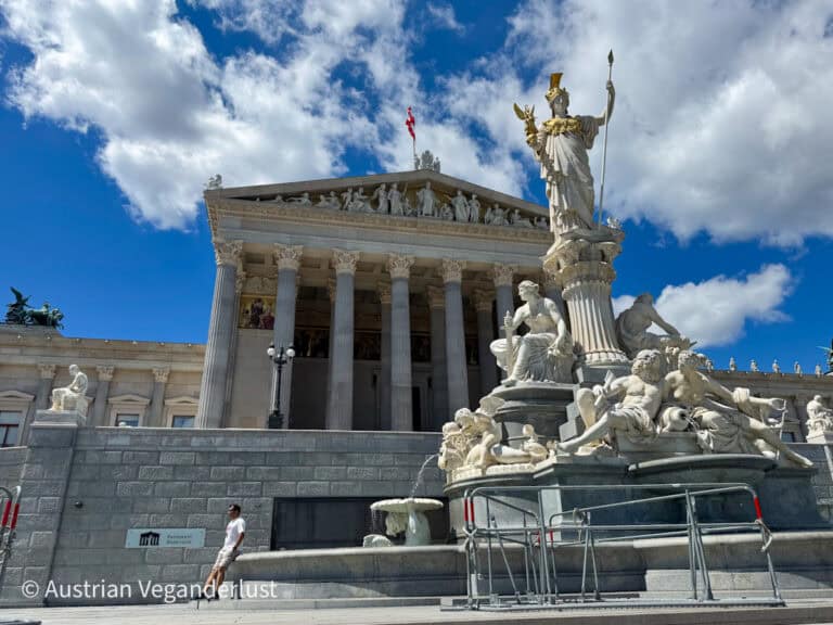 The Austrian Parliament in Vienna with its classical columned portico and sculpted pediment under a blue sky with white clouds. The Pallas Athena Fountain stands in front with the goddess in a golden helmet holding a spear and a small winged figure above reclining marble figures. A sign on the wall reads “Parlament Österreich” and a man in a white shirt leans against the stone base nearby.